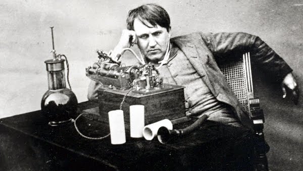 A photo of Thomas Edison, in a slouching pose behind some of his inventions.